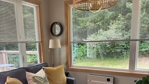 A living room with a grey sofa, a round coffee table, a woven pendant light, and a view of greenery through large windows.