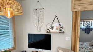 A living room with a TV, macrame wall decor, a wooden shelf with decorative items, a sofa, and a patterned rug.