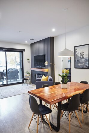 Ein moderner Essbereich mit einem Holztisch und -stühlen, einem an der Wand befestigten Fernseher und einer Glasschiebetür, die auf eine Terrasse im Freien führt.