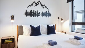 Un letto rifatto con cuscini, un comodino con libri, un'opera d'arte raffigurante montagne e alberi, e una finestra con vista.
