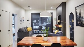 Un interno moderno con un tavolo da pranzo in legno, sedie nere, un divano grigio con cuscini gialli, una lampada a sospensione e un'ampia finestra con vista su alberi e un edificio.