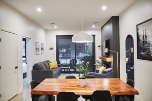 Ein modernes Interieur mit einem Holzesstisch, schwarzen Stühlen, einem grauen Sofa mit gelben Kissen, einer hängenden Pendelleuchte und einem großen Fenster mit Blick auf Bäume und ein Gebäude.