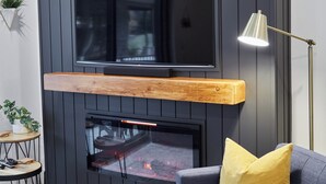 Un soggiorno moderno con una TV a schermo piatto montata su una parete di legno scuro, un caminetto sottostante, un divano blu con un cuscino giallo, una lampada da terra e un piccolo tavolino con una pianta.