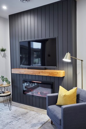 Ein modernes Wohnzimmer mit einem an einer dunklen Holzwand befestigten Flachbildfernseher, einem Kamin darunter, einem blauen Sofa mit einem gelben Kissen, einer Stehlampe und einem kleinen runden Tisch mit einer Pflanze.