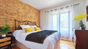 A bedroom with a brick wall, a bed with white bedding and yellow pillows, a wooden dresser, and a window with sheer curtains. - House in La Cerdanya, Ger. Surrounded by nature (La Pleta de Saga)