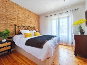 Ein Schlafzimmer mit Backsteinwand, einem Bett mit weißer Bettwäsche und gelben Kissen, einer Holzkommode und einem Fenster mit transparenten Vorhängen.