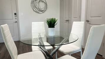 A glass-top dining table with four white chairs, a potted plant, and a wall-mounted abstract art piece.