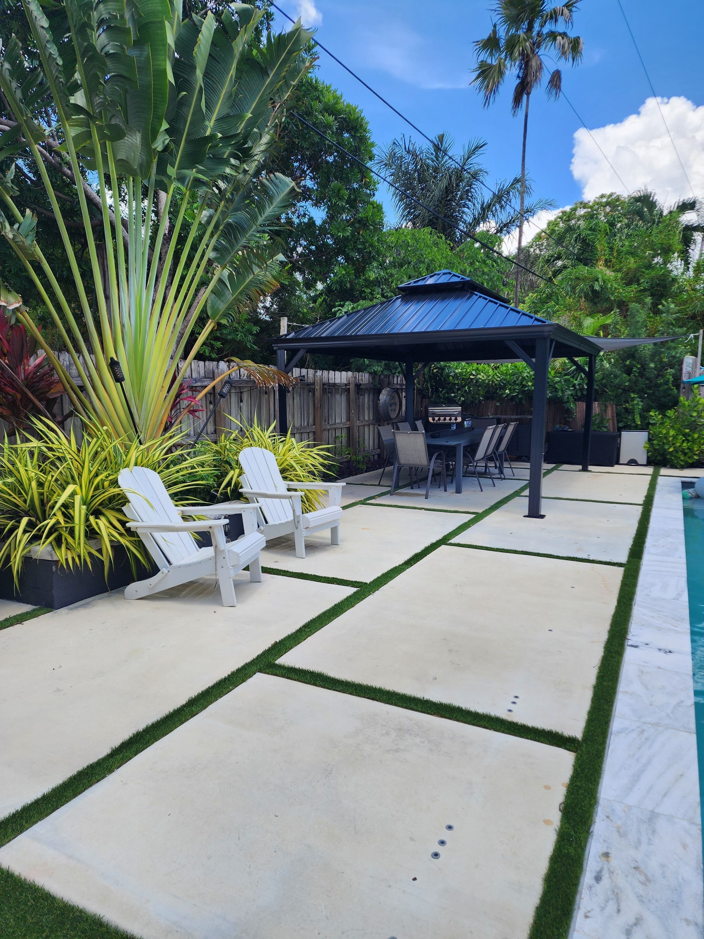 A backyard with a pool, a covered barbecue area, and white Adirondack chairs.