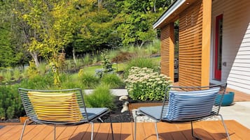 En terrasse med to stole og et lille bord, omgivet af en have med forskellige planter og en trækonstruktion i baggrunden.