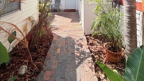 A narrow brick pathway leading to a white door, surrounded by potted plants and a palm tree. - Private Beach Cottage Compound • Steps to Sand (Indian Rocks Beach)
