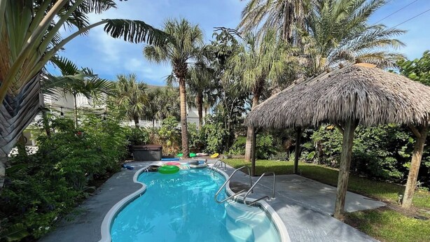 A swimming pool with a thatched roof structure, surrounded by palm trees and a paved walkway. - Private Beach Cottage Compound • Steps to Sand (Indian Rocks Beach)