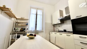 A modern kitchen with white cabinetry, a central island with a bowl of fruit, and a window with curtains. - Alex's House La Spezia Migliarina 10 min from Migliarina Station (La Spezia)