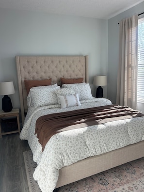 A bedroom with a tufted headboard, floral bedding, a brown throw blanket, two bedside lamps, and a window with curtains. - Modern Condo Near Wright-Patt, Shops & Dining (Beavercreek)