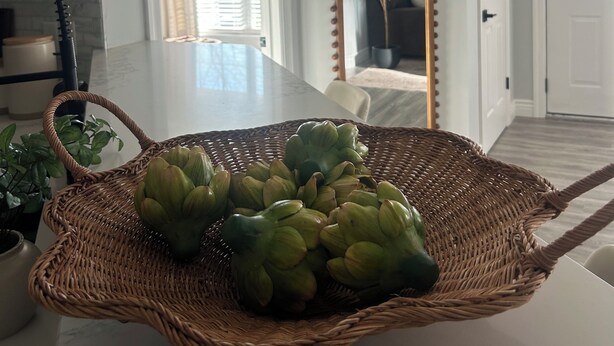 Un comedor con una mesa, sillas, una cesta de mimbre con frutas verdes, un espejo y una planta en maceta.