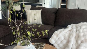 Una sala de estar con un sofá marrón oscuro, una manta mullida blanca, un jarrón blanco con ramas y una mesa de centro de madera.