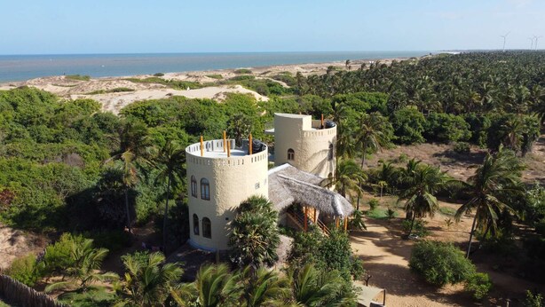 A coastal building with two towers, surrounded by dense palm trees and a beach in the background.