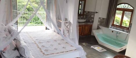 A bedroom with a bed, a bathroom with a bathtub, and a view of greenery.
