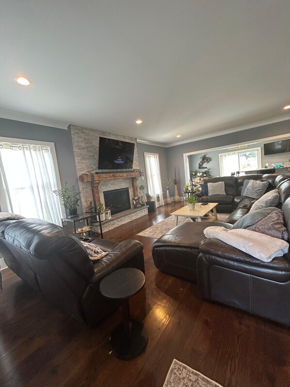A living room with a fireplace, leather sofas, a television, and a stone accent wall. - Luxury Coastal Retreat ✨ 5 min to Beach,Sleeps 10,BBQ + Patio (Long Branch)