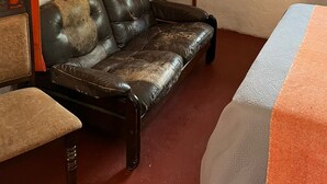 Huoneessa on nahkasohva, puinen hylly, jossa on esineitä, ikkuna, jossa on verhot, ja sänky, jossa on kuviollinen päiväpeite.