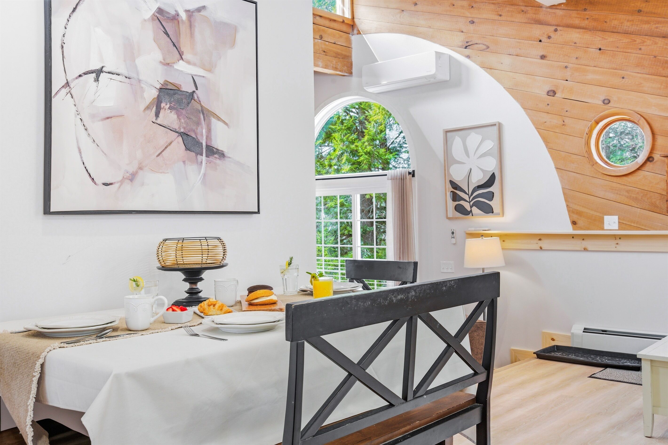 A dining area with a table set for two, a framed abstract painting, and a window with a view of greenery.