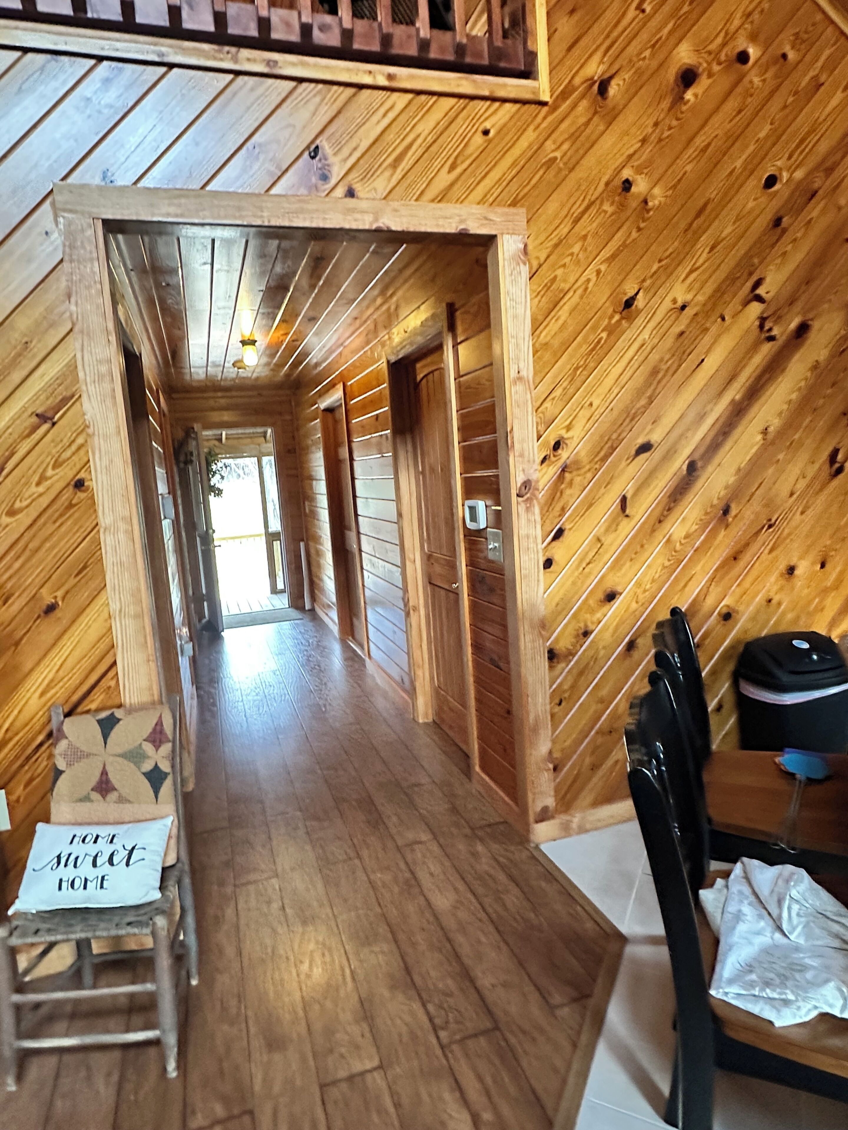Een houten interieur met een lange gang die naar een deur leidt, een kleine tafel met een stoel, en een bordje waarop staat 'HOME sweet HOME'.