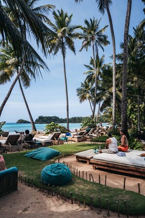 A beachside lounge area with lounge chairs and a wooden table, surrounded by palm trees and a view of the ocean. - 2-bed Pool View on Beach | Sol de Arena C1 (Las Terrenas)