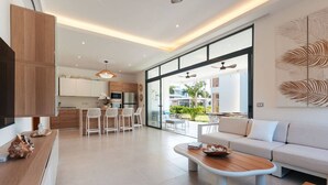 Una sala moderna con un sofá, una mesa de centro y taburetes de bar. Se observa una cocina al fondo con un ventanal grande.