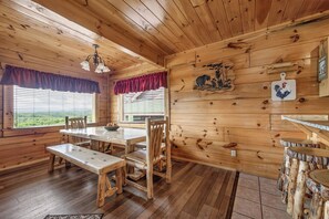Interior de uma cabana de madeira com uma mesa de jantar, bancos, um candelabro e uma janela com persianas.