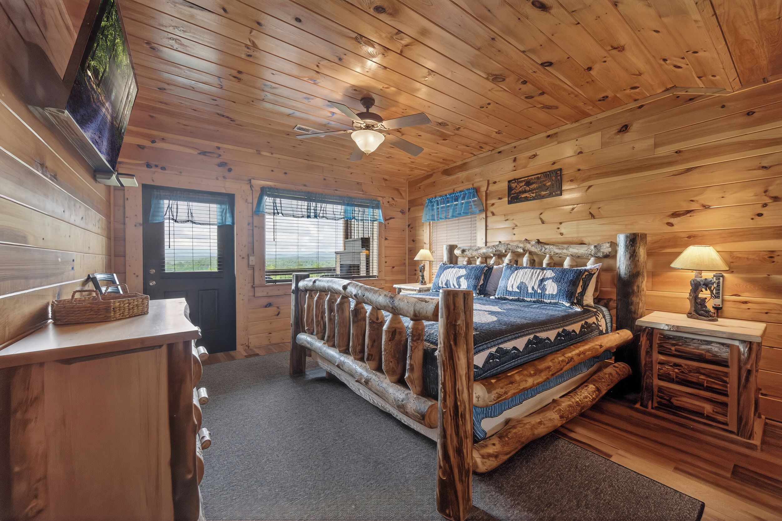A wooden cabin bedroom with a bed, a desk, a chair, a ceiling fan, and a TV mounted on the wall.