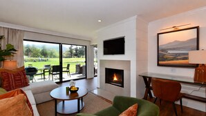 Ein modernes Wohnzimmer mit Kamin, einem an der Wand befestigten Fernseher und Blick auf eine grüne Landschaft durch große Fenster.