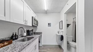 Una cocina compacta con armarios blancos, encimera de granito, microondas y lavamanos.
