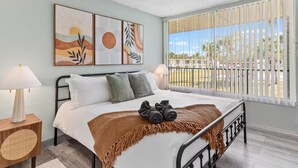 Un dormitorio con cama, almohadas, manta, lámpara, mesita de noche y cuadros en la pared.