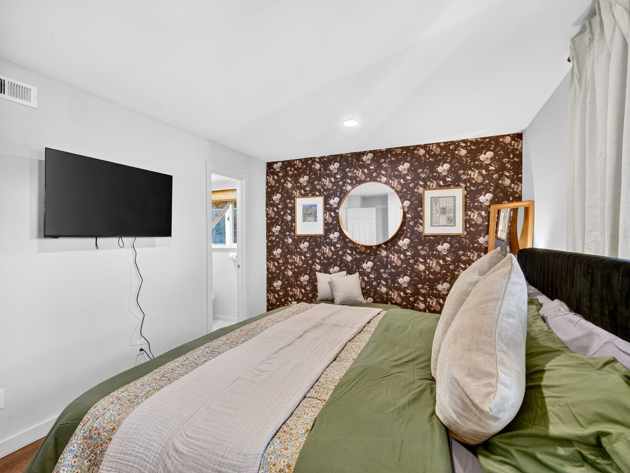 A bedroom with a bed, a flat-screen TV mounted on the wall, a window with a view, and a door leading to another room.