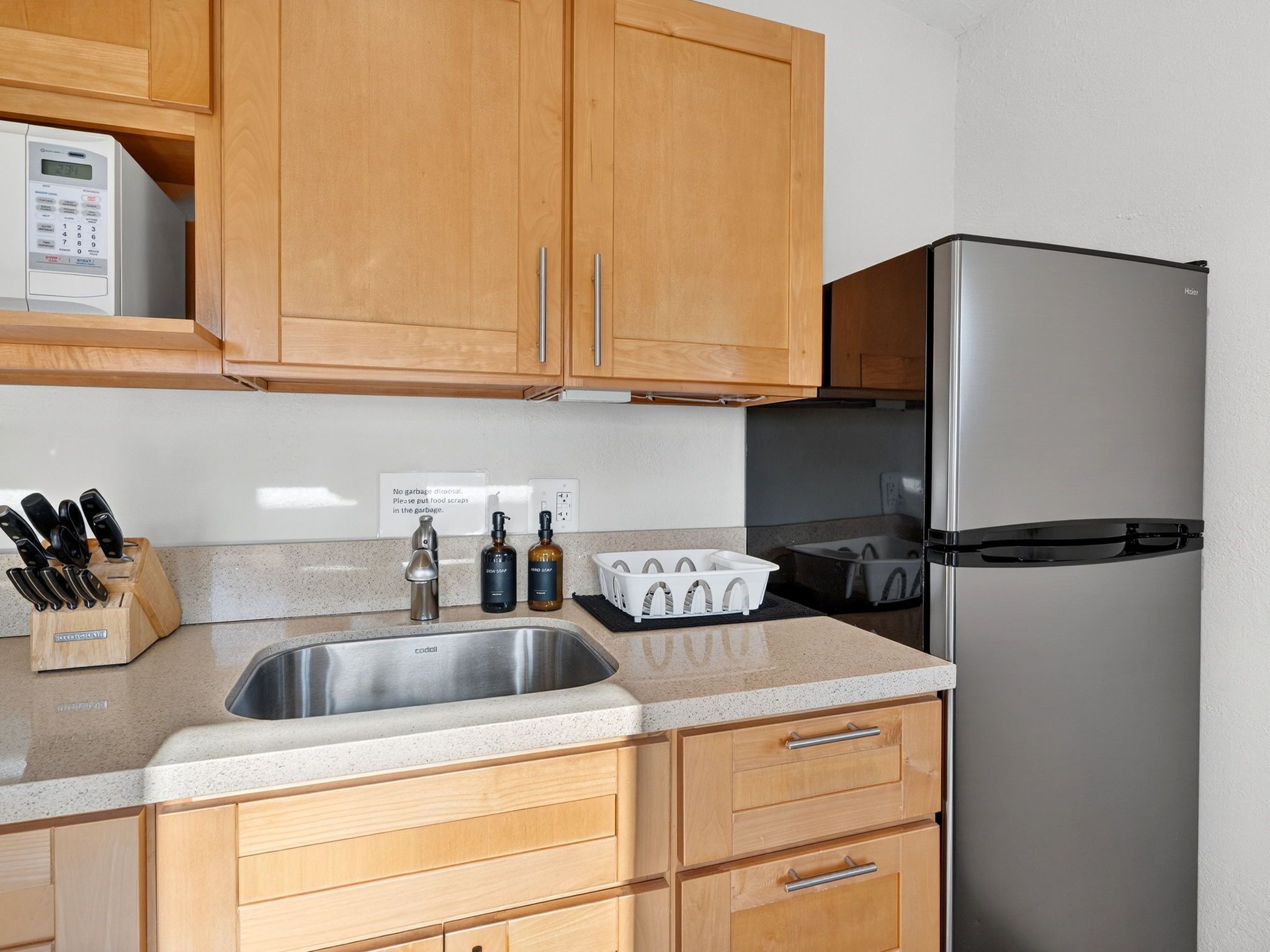 A kitchen with wooden cabinets, a stainless steel sink, a microwave, a refrigerator, and a knife block.