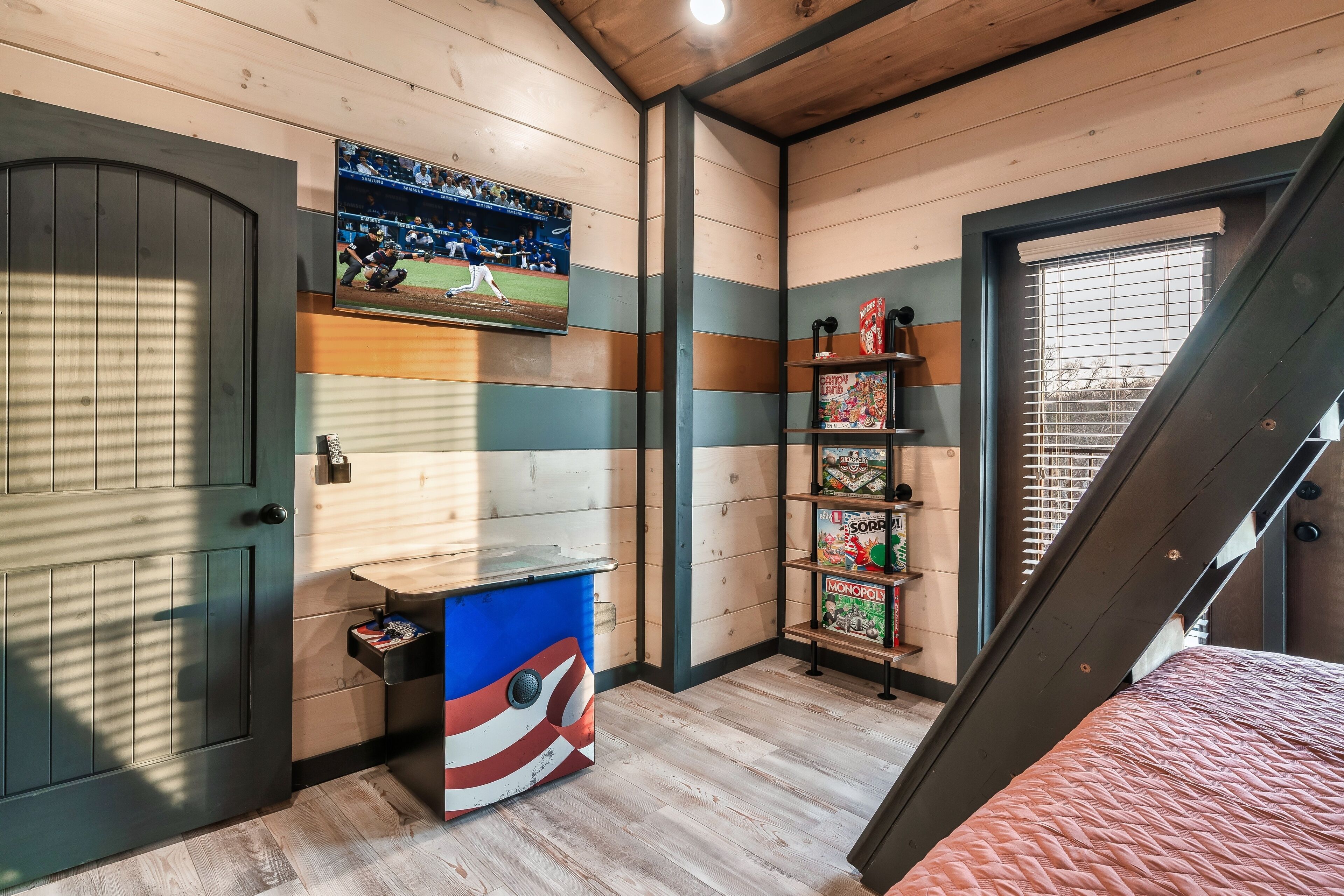A room with a wooden ceiling, a television mounted on the wall, a shelf with board games, and a door.