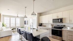 A modern kitchen with a central island, stainless steel appliances, and a dining area with a white sofa and bar stools. - Modern 1200sf 2bed apto in Frisco (Frisco)