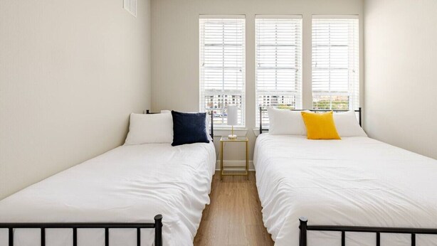 A room with two beds, white bedding, a small bedside table with a lamp, and a window with blinds. - Modern 1200sf 2bed apto in Frisco (Frisco)