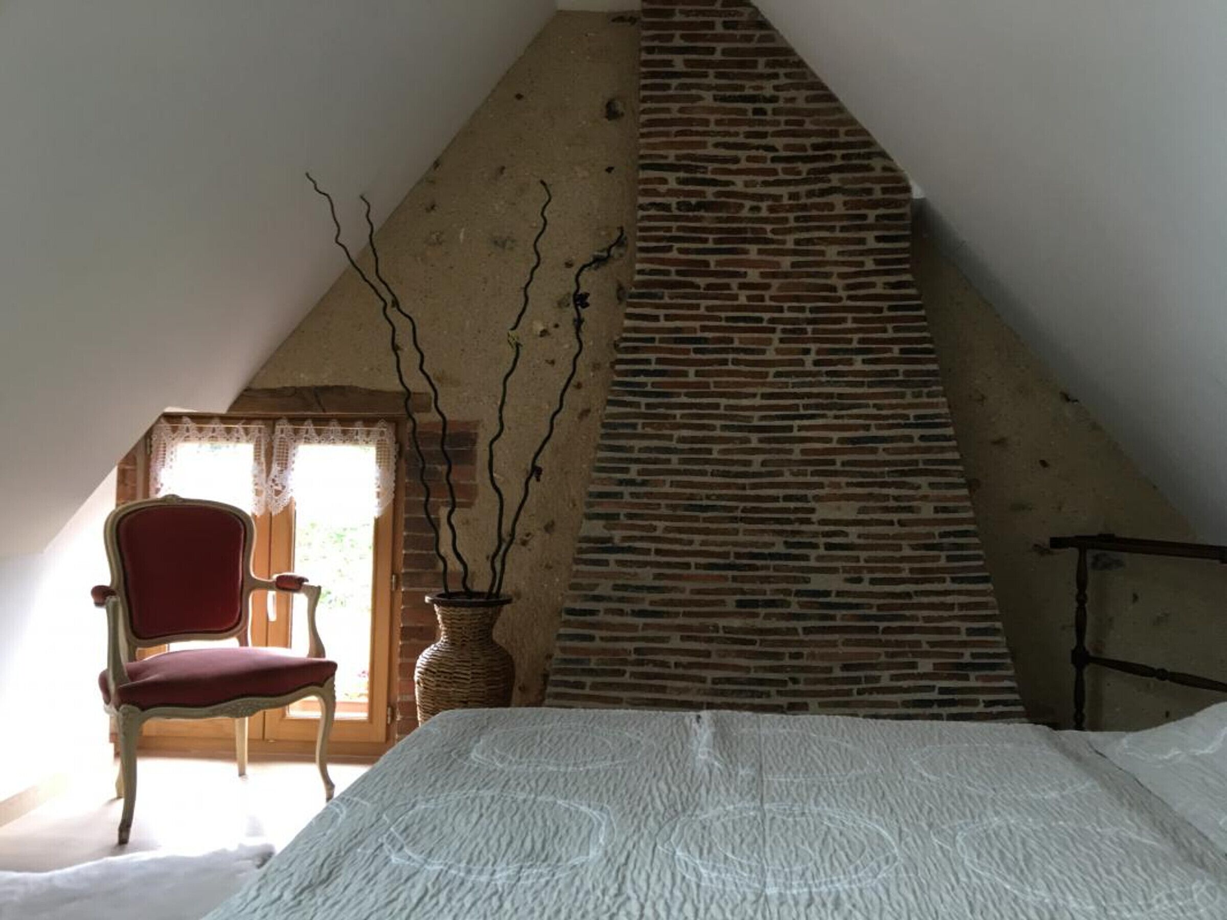A room with a sloped ceiling, a brick fireplace, a red chair, and a vase with branches.