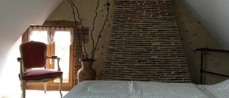 A room with a sloped ceiling, a brick fireplace, a red chair, and a vase with branches.
