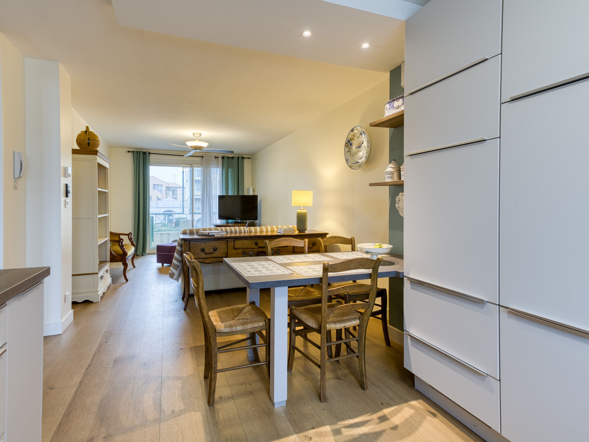 A modern kitchen with a dining area, wooden chairs, a table with papers, a television, and a window with curtains.