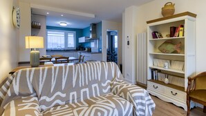 A living room with a patterned sofa, a bookshelf with decorative items, and a view into the kitchen. - Apartmentby the sea - 4 guests (Châtelaillon-Plage)