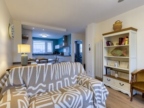 A living room with a patterned sofa, a bookshelf with decorative items, and a view into the kitchen. - Apartmentby the sea - 4 guests (Châtelaillon-Plage)