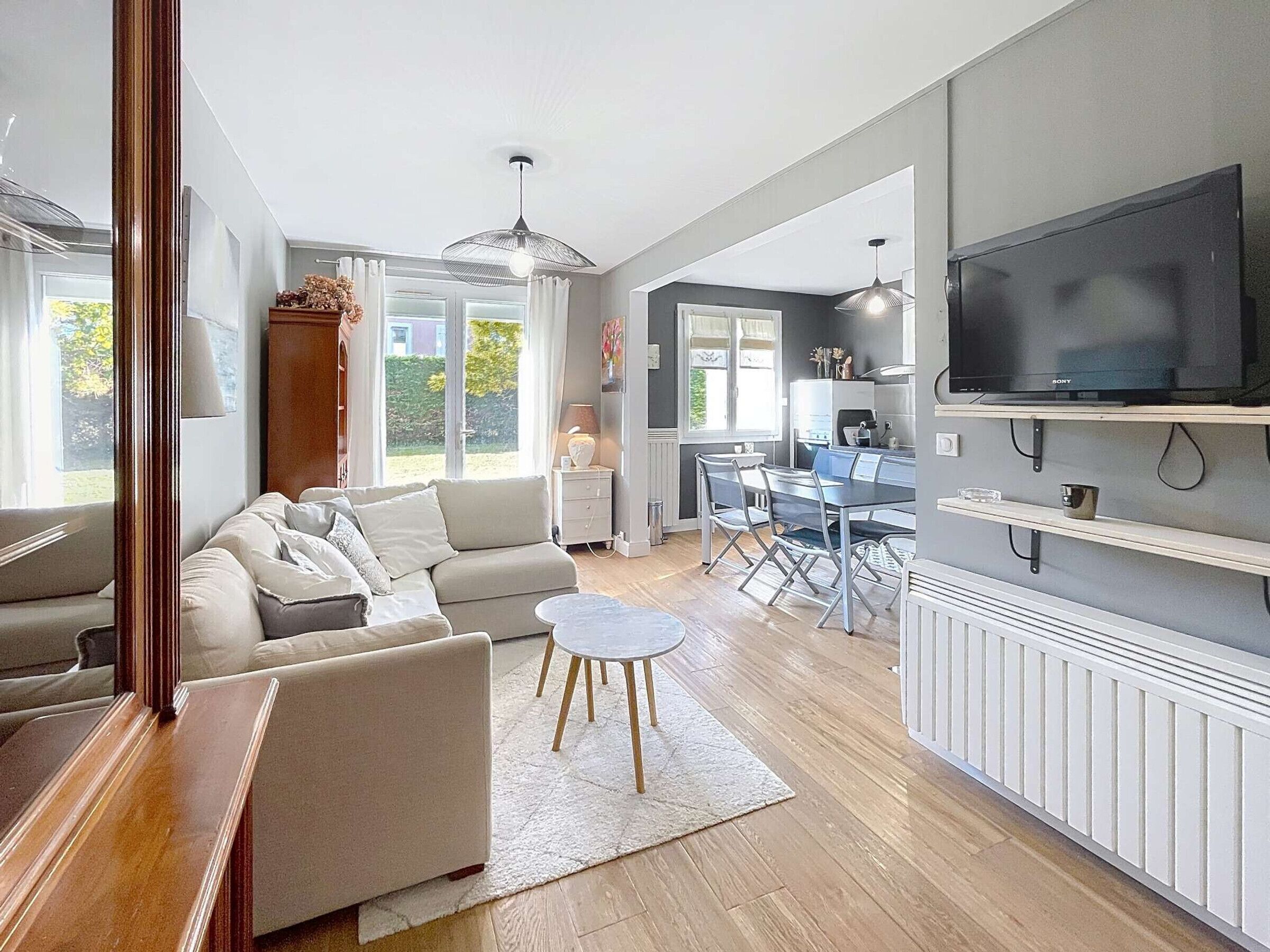 A modern living room with a sofa, a coffee table, and a dining area with chairs and a table. There is a TV mounted on the wall and a window providing natural light.