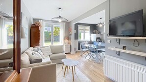 A modern living room with a sofa, a coffee table, and a dining area with chairs and a table. There is a TV mounted on the wall and a window providing natural light. - Ground floor apartment, near the sea (Jullouville)
