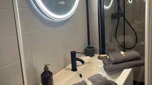 Une salle de bain avec un miroir rond, un lavabo, une douchette à main et une paroi de douche en verre.