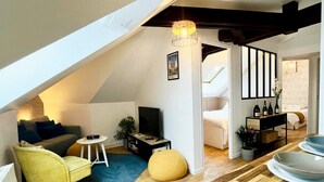 Un appartement de style « loft » avec un plancher en bois, un canapé, un fauteuil jaune, une petite table ronde, un téléviseur et un coin repas avec des assiettes et des bouteilles.