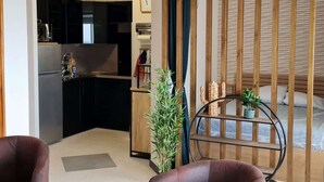 Un espace de vie compact comprenant un coin cuisine, un lit avec une tête de lit en bois, un petit coin salon et une plante en pot.