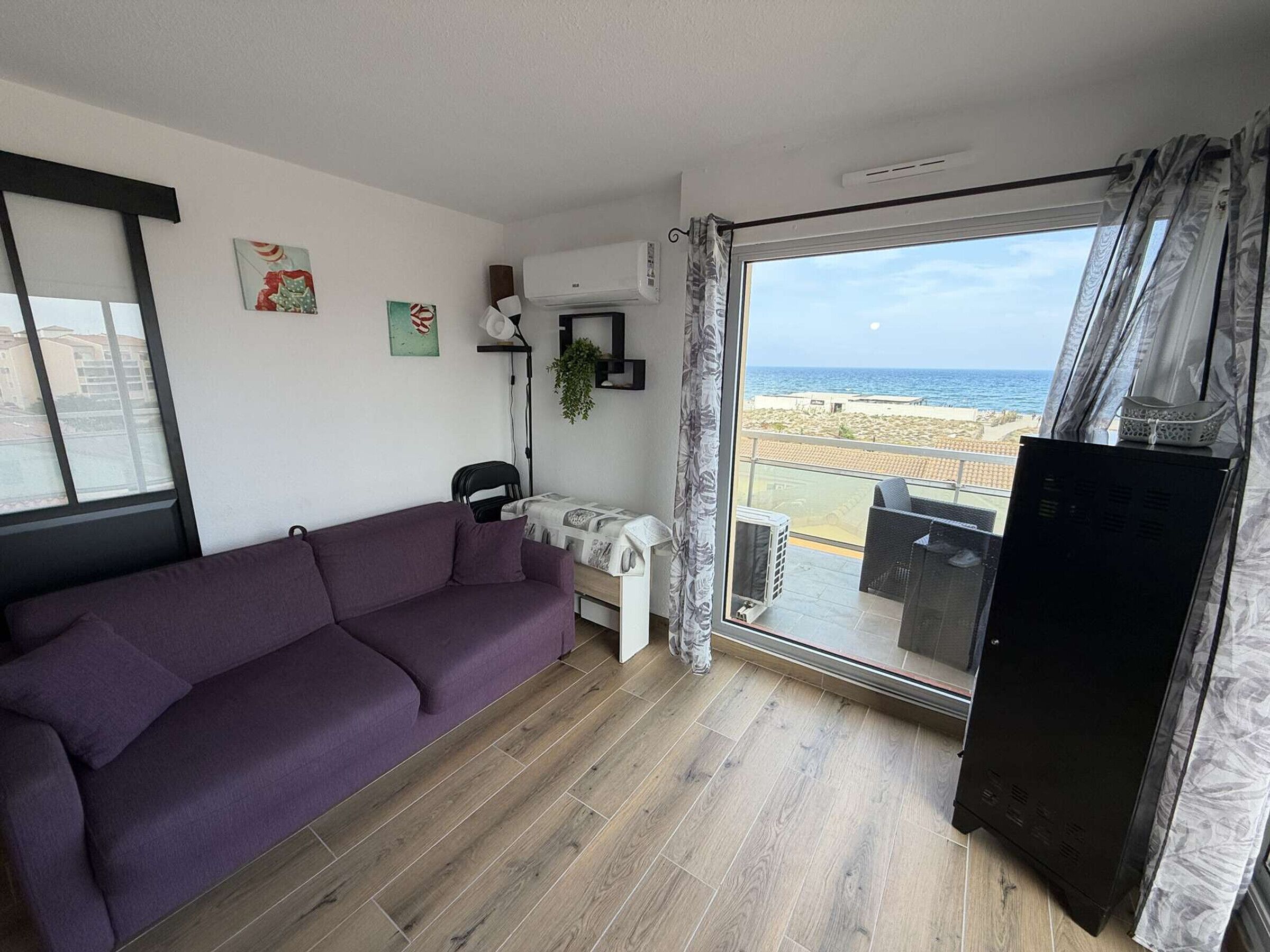 A living room with a purple sofa, a small table, a TV stand, and a large sliding glass door leading to a balcony with a view of the beach.