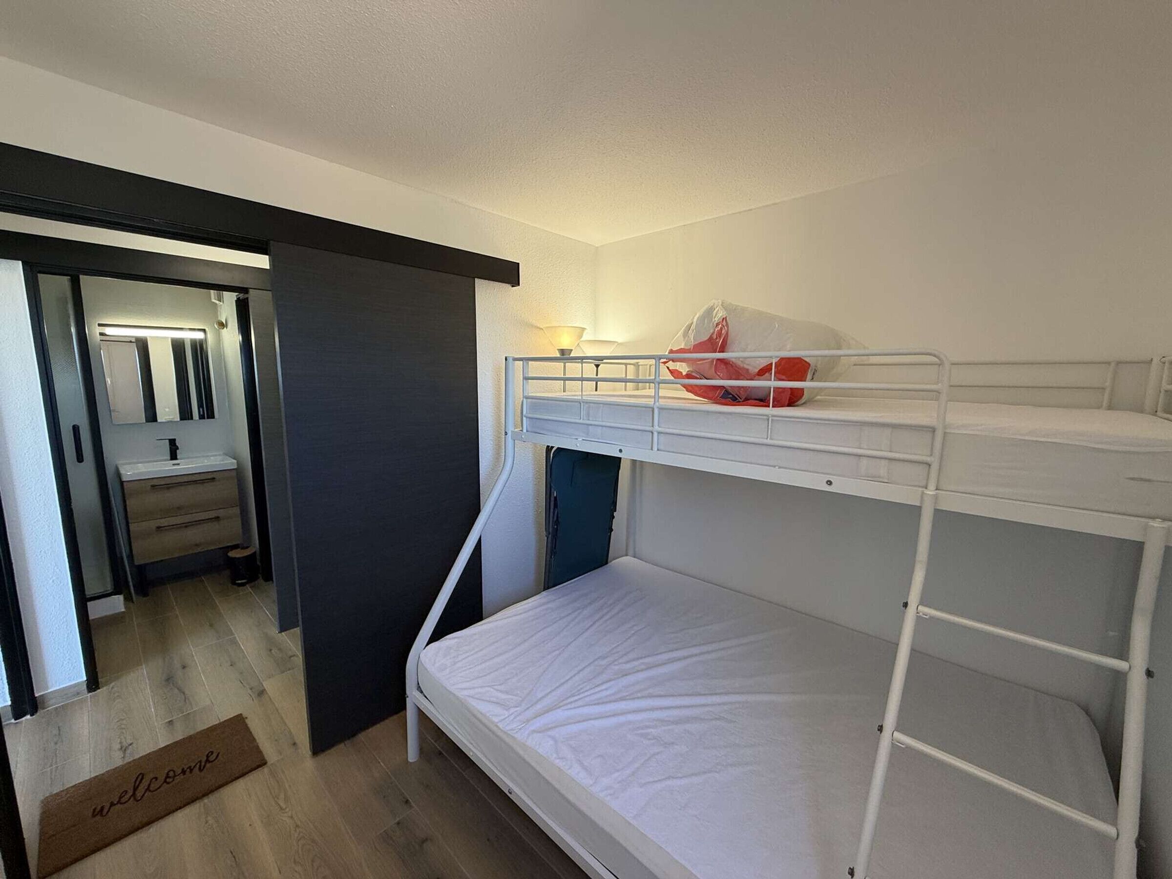 A bunk bed with a white mattress, a small bedside table, and a lamp. There is a door leading to a bathroom with a mirror and a sink.
