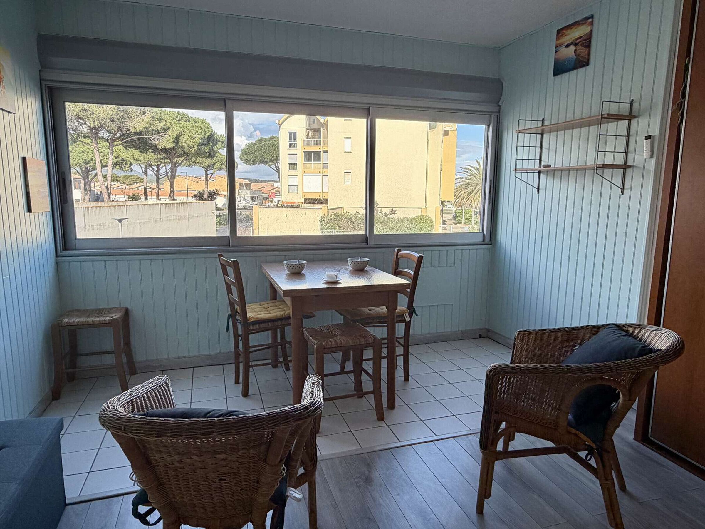 A dining area with a table and chairs, a window with a view of buildings, and a shelf with items.